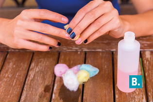 Fotografia de uma pessoa sentada diante de uma mesa retirando esmalte das unhas usando um pedaço de algodão. Sobre a mesa de madeira, há vários pedaços de algodão coloridos em tons de rosa, azul, amarelo e branco. À direita, há um pequeno frasco de plástico transparente contendo um líquido rosa, e indicado pela letra B.