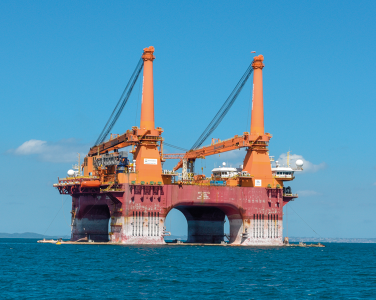 Fotografia de uma plataforma de petróleo no mar. A plataforma é grande, de cor predominantemente laranja e possui duas torres altas. Há diversas estruturas e equipamentos visíveis na plataforma, incluindo guindastes e antenas. A base da plataforma está submersa na água azul do mar.