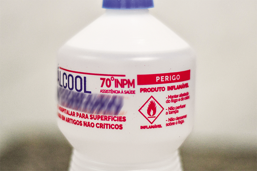 Fotografia de um frasco de álcool com rótulo predominante em branco, azul e vermelho. O rótulo contém as palavras Álcool em letras grandes e 70 graus I N P M em vermelho. Há a descrição Perigo e o aviso de Produto inflamável, acompanhados de um símbolo com uma chama em vermelho.