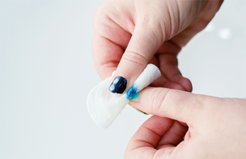 Fotografia de uma mão segurando um pedaço de algodão branco que está sendo usado para remover esmalte azul de uma unha.