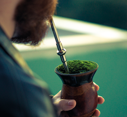 Fotografia de uma pessoa segurando uma cuia de chimarrão com erva-mate verde preenchendo o recipiente. A pessoa está bebendo o chimarrão com um canudo metálico que está inserido na cuia.