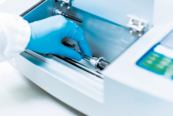 Fotografia de um equipamento de laboratório sendo manipulado. Uma mão com luva azul está manuseando uma amostra em uma cuba interna do equipamento, que é metálico e possui tampas articuladas.