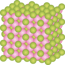 Ilustração de uma estrutura representada por esferas verdes e esferas rosas organizadas em camadas alternadas e que juntas compõe o formato de um bloco retangular. As esferas estão dispostas de forma regular e simétrica, formando um padrão repetitivo, na qual as esferas verdes estão posicionadas principalmente nas camadas superiores e nas bordas, e as esferas rosas preenchem a parte interna e se conectam às verdes em todos os lados.