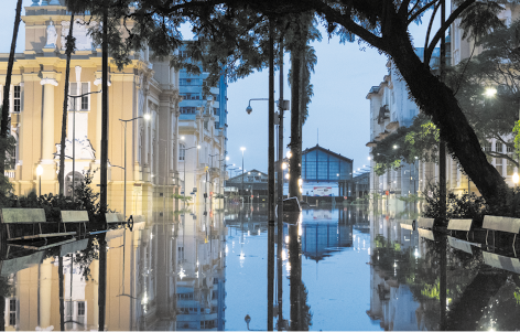 Fotografia de um ambiente urbano com uma rua alagada refletindo os prédios e árvores ao redor. Nas laterais há vários prédios e árvores com galhos e folhas estão distribuídas em primeiro plano, com uma delas em destaque, inclinada para o meio da rua.