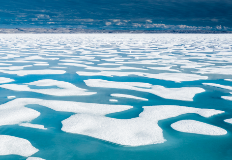 Fotografia de uma paisagem com uma extensa camada de gelo branco com áreas derretidas, formando manchas irregulares de água azul.