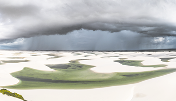 Fotografia de uma paisagem com dunas de areia branca intercaladas por lagoas verdes de diferentes tamanhos. O céu está coberto por nuvens densas e escuras.