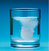 Fotografia de um copo de vidro transparente contendo água, com um comprimido efervescente em processo de dissolução. Pequenas bolhas brancas e espuma se formam ao redor do comprimido, subindo em direção à superfície da água.
