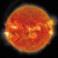 Fotografia do Sol com cores em tons de vermelho, laranja e amarelo, destacando áreas brilhantes. Há explosões de luz nas bordas e detalhes granulados na superfície.
