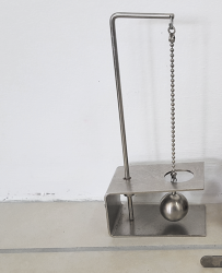 Fotografia de um aparelho metálico. Ele é composto por uma estrutura com base retangular, um suporte vertical e uma corrente metálica pendurada no topo do suporte. Na extremidade inferior da corrente, a qual passa por dentro de uma abertura circular há uma esfera metálica.