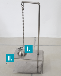 Fotografia de um aparelho metálico composto por uma base retangular, um suporte vertical e uma corrente com uma esfera metálica pendurada na extremidade inferior. A esfera está indicada por 1, e ela está presa a uma abertura circular na base que está indicada por 2.