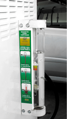 Fotografia de um equipamento para combustível em um posto de gasolina. O aparelho é composto por um tubo transparente vertical preenchido com líquido, contendo uma flutuante com marcações coloridas que indicam diferentes densidades. Há um adesivo verde ao lado do tubo com orientações e ilustrações.