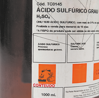 Fotografia de um rótulo de frasco contendo ácido sulfúrico. No topo do rótulo, está escrito Ácido Sulfúrico Grau H índice 2 S O índice 4. Abaixo, informações adicionais incluem avisos como Ácido sulfúrico provoca queimaduras graves e está indicado que a venda é proibida para menores de 18 anos. O volume do conteúdo é indicado como 1000 mililitros. Há um símbolo de perigo, com um ícone laranja com uma representação de corrosão.