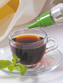 Fotografia de uma xícara de vidro transparente contendo café preto. A xícara está apoiada em um pires de vidro transparente. Próximo à xícara há uma folha verde, e acima, uma embalagem com tampa verde despeja gotas no líquido.