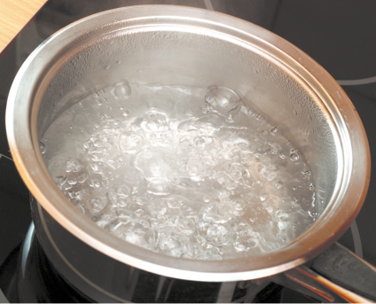 Fotografia de uma panela metálica contendo água em ebulição sobre uma superfície de aquecimento. A água está borbulhando intensamente, com bolhas visíveis na superfície e em formação no fundo da panela.