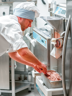 Fotografia de um homem usando uniforme branco, incluindo um chapéu branco, manuseando uma máquina de sorvete. Ele está colocando um sorvete de cor marrom em uma bandeja retangular de metal.