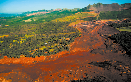 Fotografia aérea de uma paisagem com uma grande área coberta por lama em tons de marrom no centro. À esquerda, há áreas de mata preservada em tons de verde, enquanto ao fundo há colinas e montanhas que compõem o relevo.