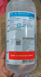 Fotografia de uma mão segurando um frasco com uma solução transparente. O rótulo do frasco exibe o texto Solução de Glicose 5 por cento, com detalhes adicionais sobre sua composição e uso médico. O frasco contém 500 mililitros de líquido, conforme indicado. Na parte inferior do rótulo, há uma faixa vermelha com o texto VENDA SOB PRESCRIÇÃO MÉDICA.