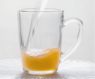 Fotografia de uma caneca de vidro transparente com alça. Dentro da caneca, há um líquido alaranjado sendo misturado com água que está sendo despejada.