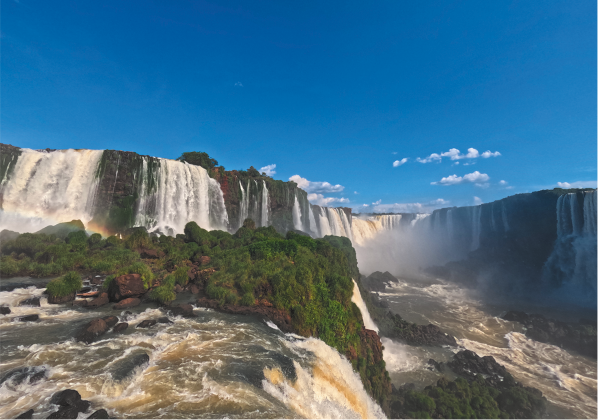 Fotografia de uma paisagem composta por um conjunto de grandes cachoeiras em um cenário ensolarado. No primeiro plano, há um corpo d’água turbulento com espuma branca formado pelas quedas d'água.