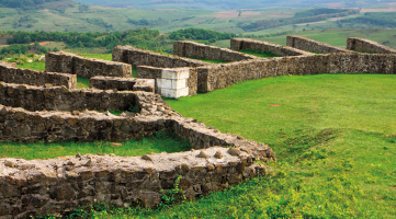 Fotografia de uma área ao ar livre com estruturas de pedra em ruínas dispostas em formações retangulares. As pedras, de coloração cinza, formam paredes baixas com um gramado verde ao redor.