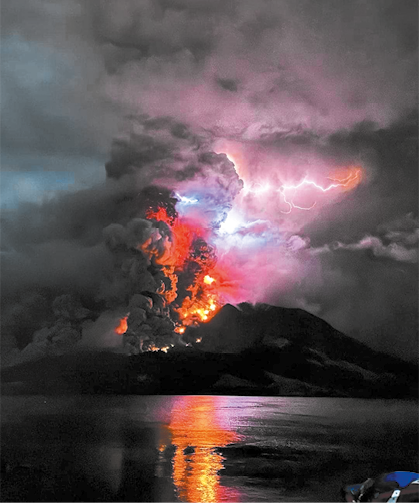 Fotografia de um vulcão em erupção durante a noite. No centro o vulcão expulsa grandes colunas de fumaça densa e cinzas escuras, que se misturam com nuvens espessas. A erupção emite luzes em tons de laranja e vermelho. Um raio brilhante atravessa as nuvens no topo.