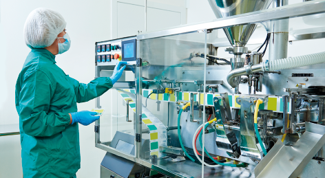 Fotografia de uma pessoa usando uniforme verde, touca branca, máscara facial e luvas, de frente para uma máquina com painel de controle eletrônico, a qual está manuseando. A máquina tem um vidro transparente e dentro dela, há vários tubos e fios.