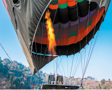 Fotografia de um balão de ar quente em processo de inflar. A estrutura do balão apresenta um padrão com faixas coloridas em preto, verde, laranja e roxo. Um jato de chama alaranjada é emitido por um queimador localizado na base do balão. Cordas conectam o balão à cesta abaixo.