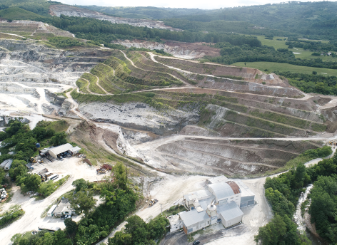 Fotografia aérea de uma área de mineração a céu aberto, evidenciando uma grande cava com cortes em terra formando patamares ao longo da encosta. A região apresenta áreas de solo exposto em tons claros. Na parte inferior, há edificações industriais, veículos e equipamentos de mineração.