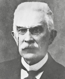 Fotografia em preto e branco de um homem idoso com cabelos curtos e claros, e bigode espesso. Ele está usando óculos e usa paletó escuro, camisa branca e uma gravata ajustada.