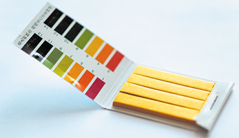 Fotografia de um kit de tiras de teste de p H, composto por várias tiras amarelas armazenadas em uma embalagem dobrável. No interior da embalagem, há uma escala de cores variando do preto ao laranja, passando por tons de verde e amarelo, associada a valores numéricos de p H.