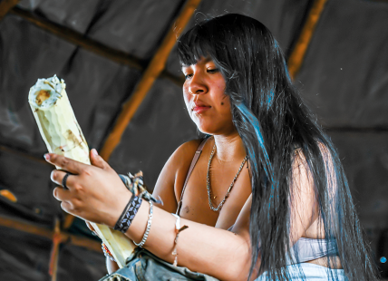 Fotografia de uma mulher com cabelos longos e escuros, segurando um pedaço de mandioca em suas mãos. Ela usa acessórios como colares, pulseiras e uma faixa no braço.