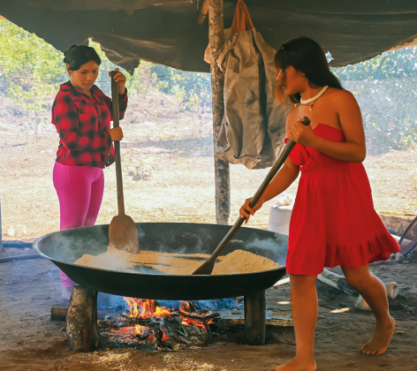 Fotografia de duas mulheres preparando alimento em uma grande panela sobre um fogo a lenha ao ar livre. A mulher à direita, usa um vestido vermelho, está com os pés descalços e está mexendo o conteúdo da panela com uma pá de madeira longa. A mulher à esquerda, usa uma camisa xadrez vermelha e calça rosa, e observa a atividade segurando uma pá semelhante.