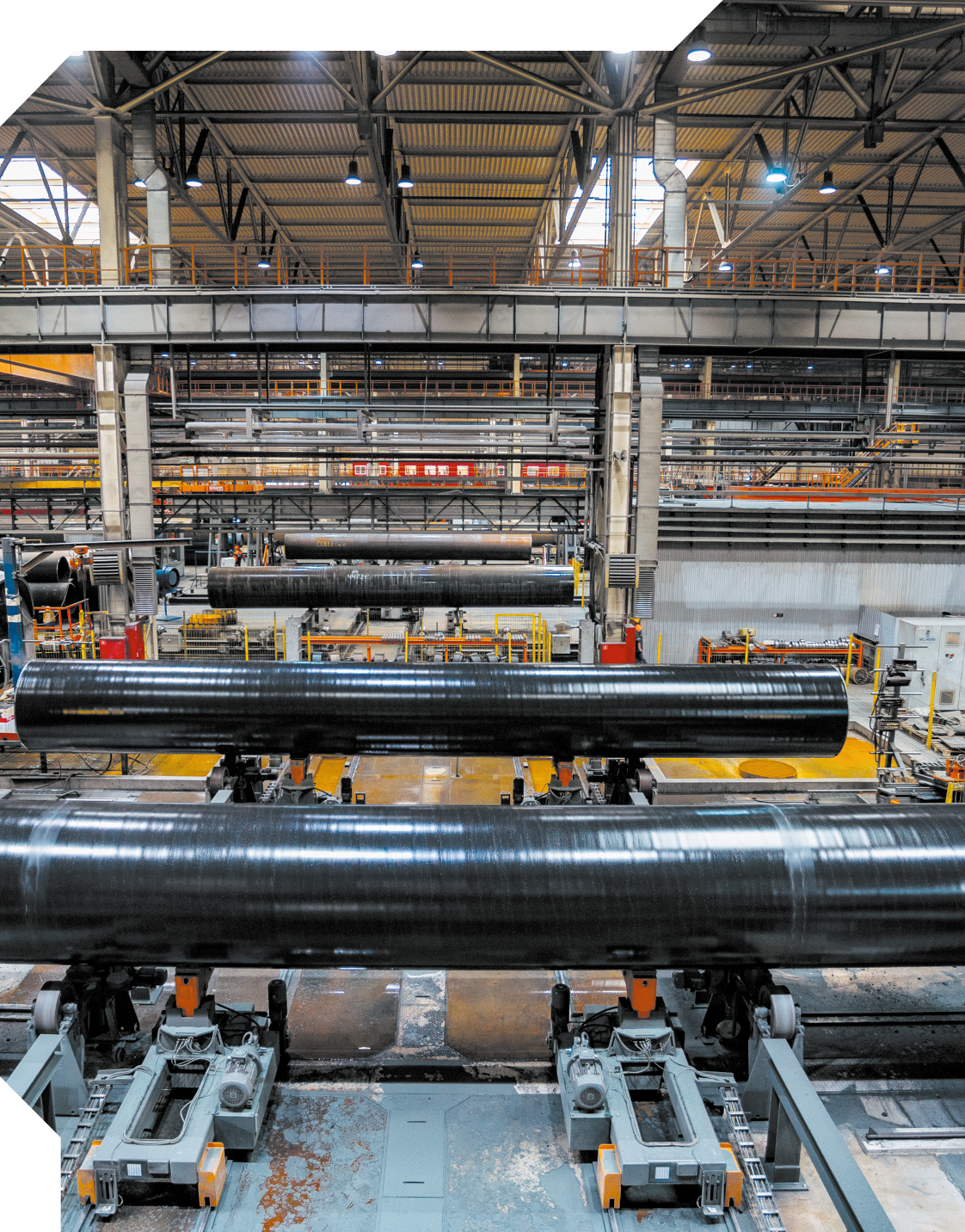 Fotografia de um ambiente industrial. Em primeiro plano, há tubos grandes e longos, de coloração preta, dispostos horizontalmente sobre um sistema de transporte. Os tubos estão apoiados em suportes metálicos. Ao fundo, há diversas prateleiras e estruturas metálicas.