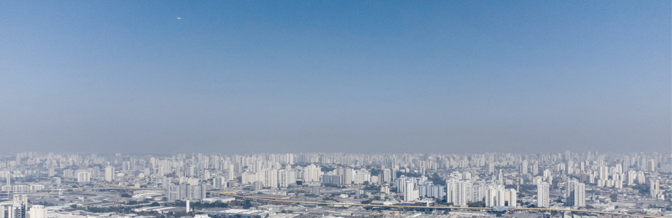 Fotografia de uma vista panorâmica de uma grande área urbana com uma vasta extensão de edifícios visto durante o dia. O céu está azul claro, com algumas áreas apresentando manchas brancas, com um dia com neblina.