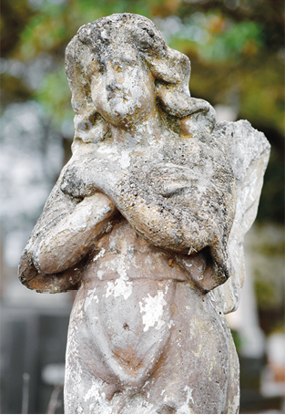 Fotografia de uma escultura parcialmente coberta por manchas escuras de desgaste e mofo. A figura tem cabelos ondulados e está com as mãos cruzadas sobre o peito.
