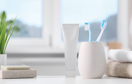 Fotografia de uma bancada de banheiro com vários itens de higiene. No centro, há um tubo de creme dental branco, ao lado de um copo branco contendo duas escovas de dentes.