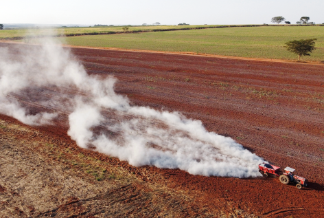 Fotografia de uma área rural, onde um trator está trabalhando em um campo de terra vermelha. O trator está soltando uma nuvem de fumaça branca enquanto se movimenta.