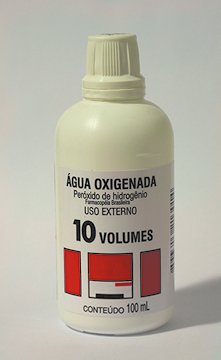 Fotografia de um frasco de plástico branco com rótulo escrito: ÁGUA OXIGENADA. Peróxido de hidrogênio e uso externo. 10 volumes. Conteúdo: 100 mililitros.