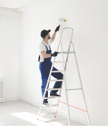 Fotografia de um homem pintando uma parede. Ele usa boné, macacão azul e luvas, e está em uma escada pintando a parede de cor branca e com o auxílio de um rolo de pintura.