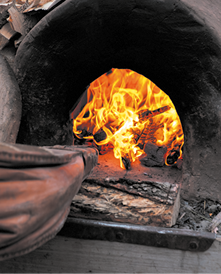 Fotografia de um forno a lenha aceso com chamas intensas em tons de amarelo e laranja consumindo pedaços de madeira. À frente, na direção do fogo está um soprador.