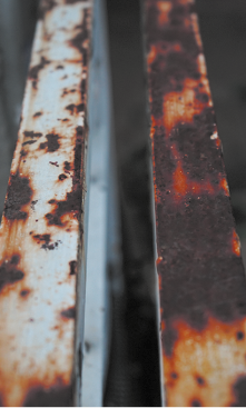 Fotografia de duas barras metálicas dispostas paralelamente, ambas com sinais de corrosão. A barra à esquerda possui uma superfície predominantemente branca com manchas de ferrugem espalhadas em tons de marrom e laranja. A barra à direita apresenta uma superfície mais escura, com áreas extensas de corrosão em tons de marrom avermelhado.