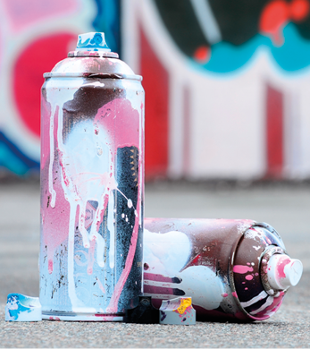 Fotografia de duas latas de spray de tinta colocadas sobre uma superfície de concreto. Uma das latas está em pé, enquanto a outra está deitada, e ambas apresentam resíduos de tinta escorrida em cores como branco, rosa e azul claro, cobrindo parcialmente sua superfície metálica.