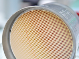Fotografia de um filtro cilíndrico metálico com uma estrutura interna composta por uma malha de pequenos canais quadrados uniformes. A superfície da malha tem uma coloração amarelada clara.