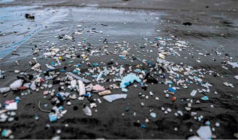 Fotografia de parte da areia de uma praia com coloração escura e com pequenos pedaços de plástico de diversas cores, tamanhos e formatos espalhados por toda parte.