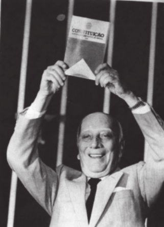 Foto de Ulysses Guimarães. Ele é um homem branco, calvo e segura no alto o texto da constituição.
