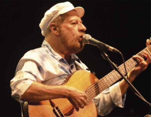 Foto de João Bosco. Ele é um homem branco, usa boina, toca violão e está cantando.