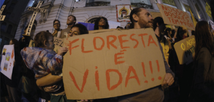 Foto de pessoas em manifestação. Elas carregam faixas e cartazes. Destaca-se um cartaz, onde está escrito: 'Floresta é vida!'.