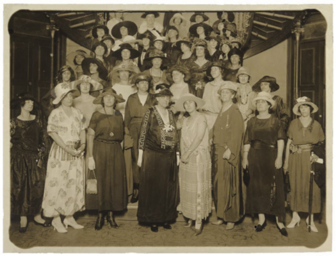 Foto de mulheres reunidas em uma escada. Elas usam vestidos longos e chapéus.