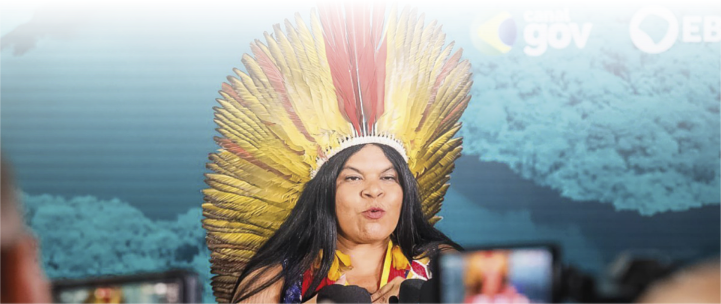 Foto de Sonia Guajajara diante da imprensa, discursando. Ela é uma mulher indígena, possui cabelos pretos e usa cocar de penas na cabeça.