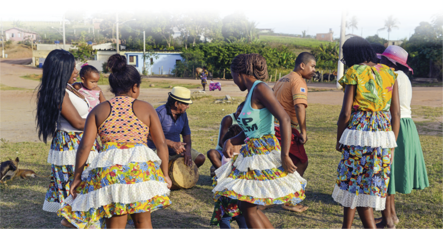 Foto de pessoas dançando juntas. As mulheres usam saias com estampas e babados. Também se destaca um homem que usa chapéu e toca tambor.